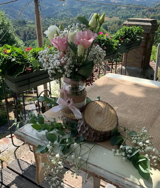 Composizione floreale su un tavolo con fiori rosa e bianchi, verde e un cesto intrecciato su un patio con vista sulle montagne.
