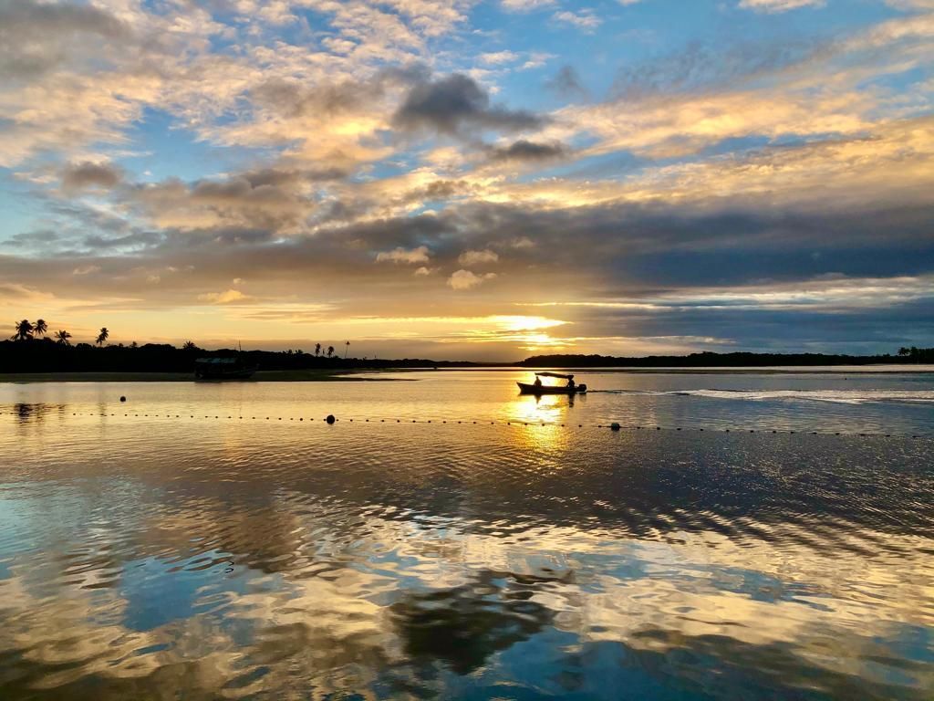 Pôr do sol sobre águas calmas, com a silhueta de um barco e nuvens douradas refletidas na superfície.