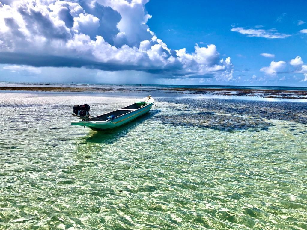 Pequeno barco verde em águas rasas e cristalinas sob um céu azul brilhante com nuvens dramáticas.