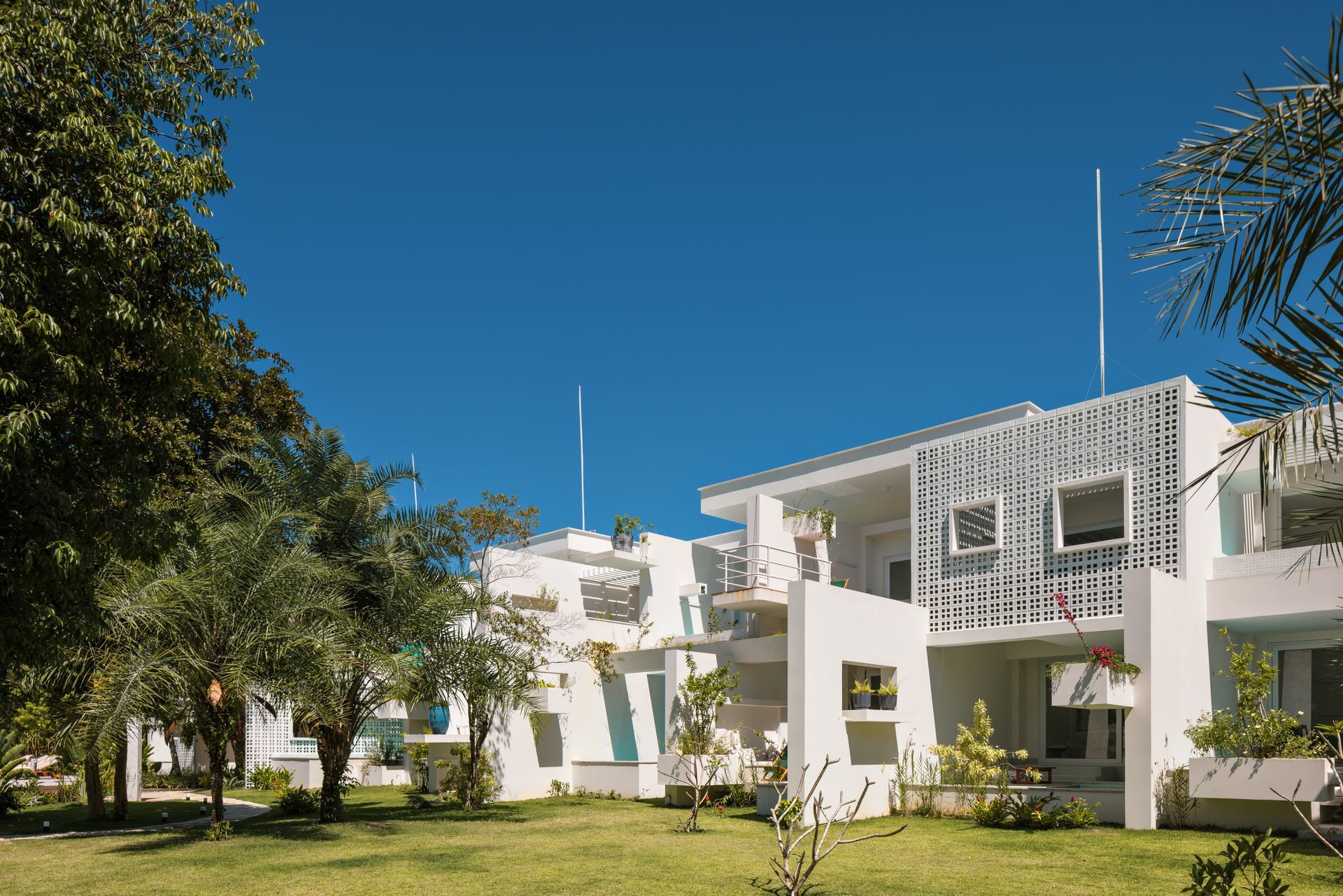 Prédio moderno branco com varandas e árvores em um gramado verde ensolarado sob um céu azul claro.