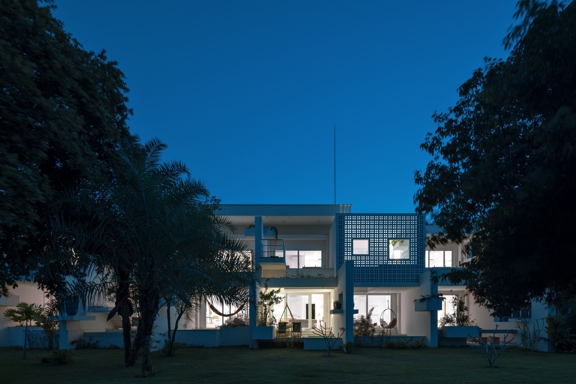 Casa moderna de dois andares, branca, ao entardecer, com janelas iluminadas brilhando sob um céu azul.