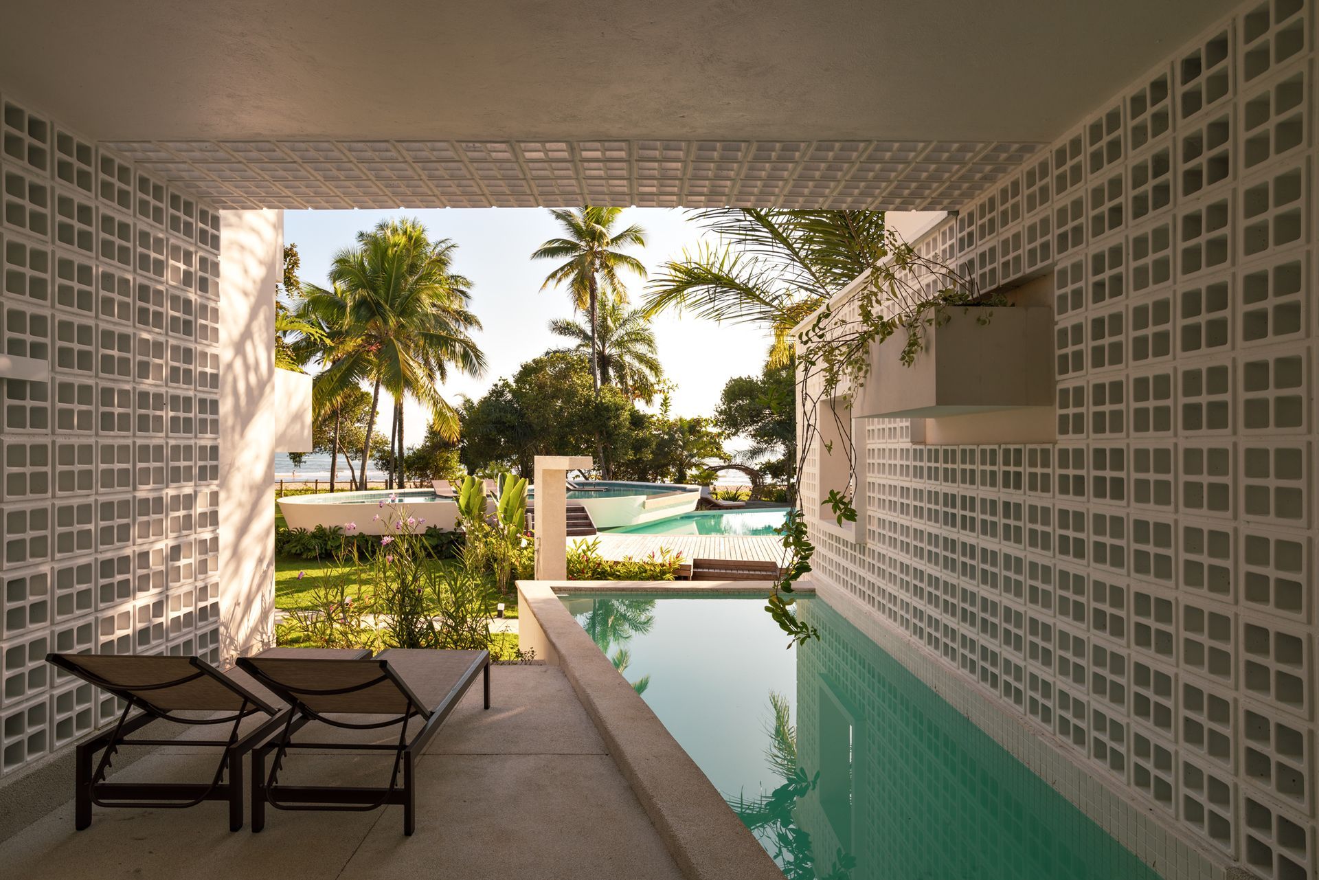 Pátio coberto com espreguiçadeiras e piscina turquesa com vista para palmeiras e um resort tropical.