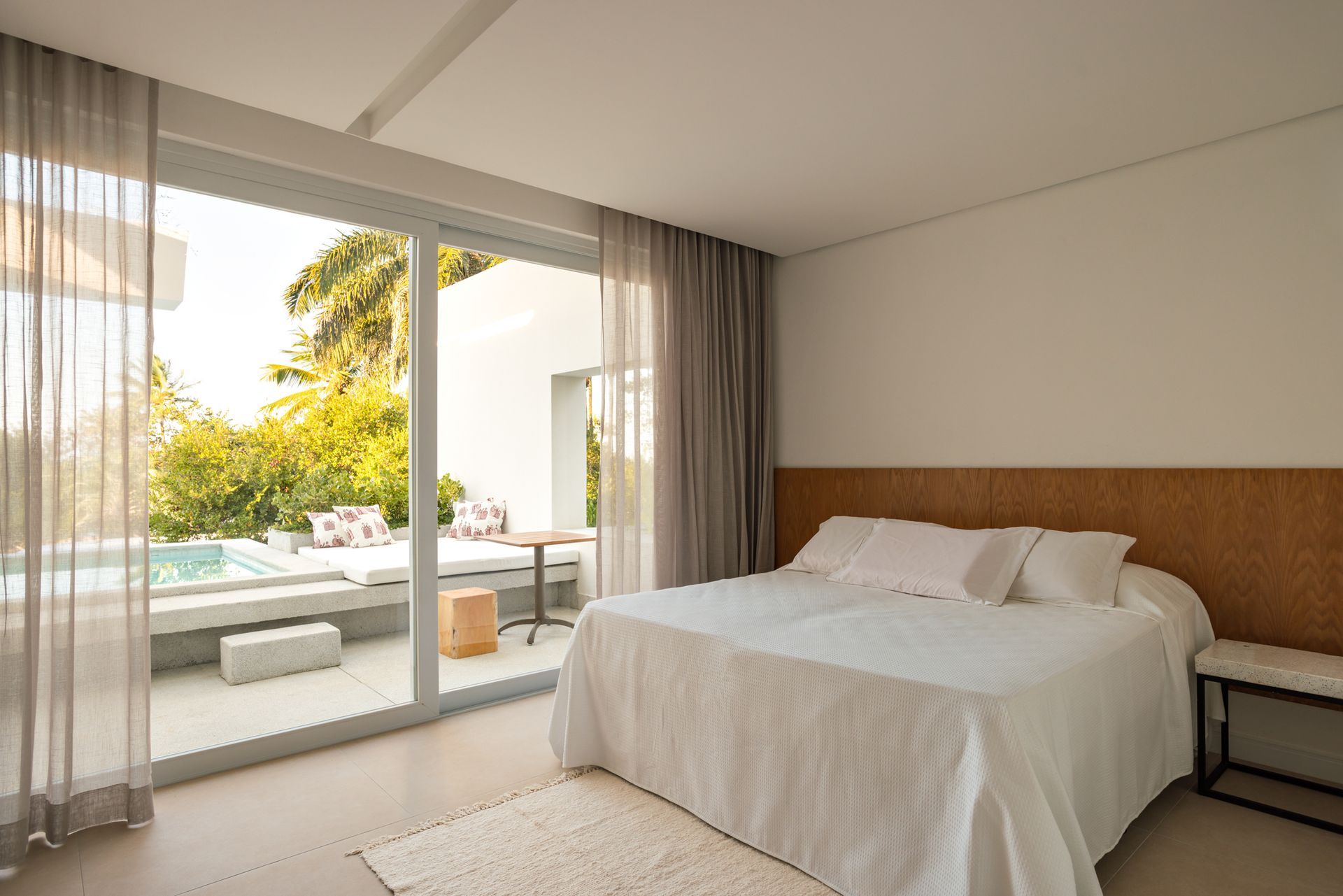 Quarto luminoso com cama ao lado de portas de vidro deslizantes que se abrem para um pátio ensolarado e área verde.