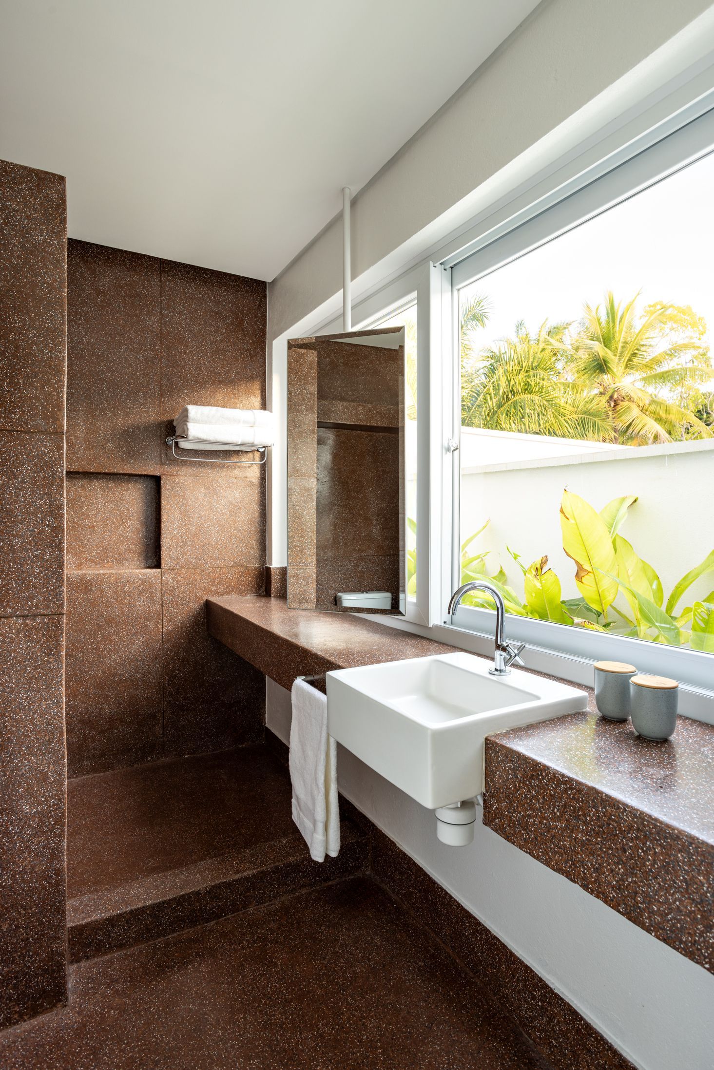 Banheiro moderno com azulejos marrons, pia suspensa, espelho e janela ampla com vista para a vegetação externa.