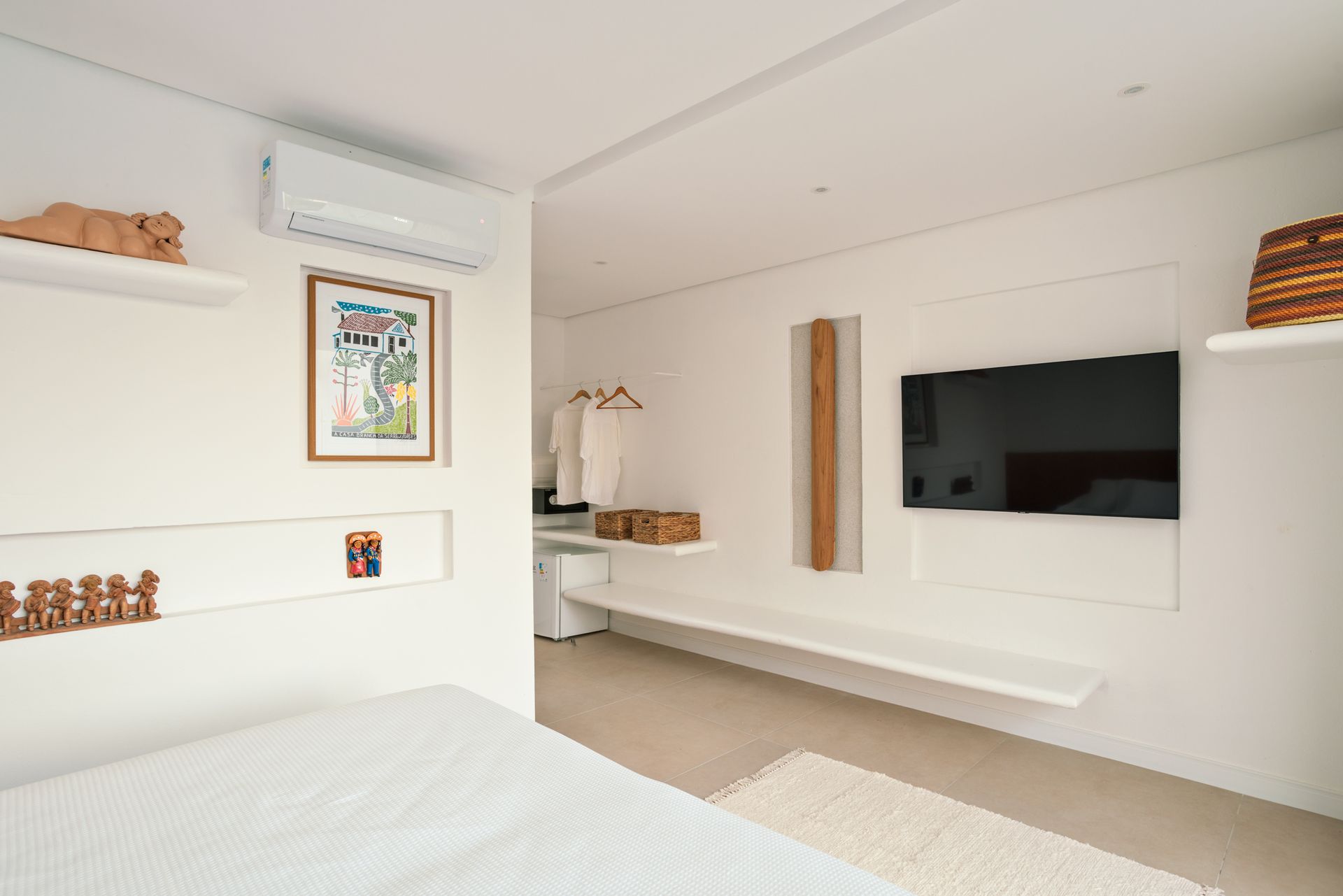 Quarto minimalista e luminoso com TV de parede, prateleiras brancas e carpete bege.