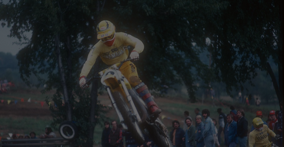 Motorcycle racer in yellow jumps over a dirt hill, crowd watches, trees and overcast sky in background.