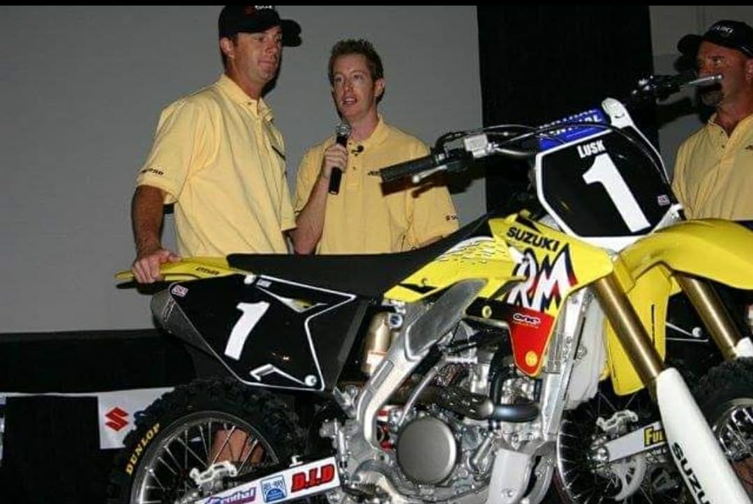 Two men in yellow shirts next to a yellow and black Suzuki motorcycle, one holding a microphone.