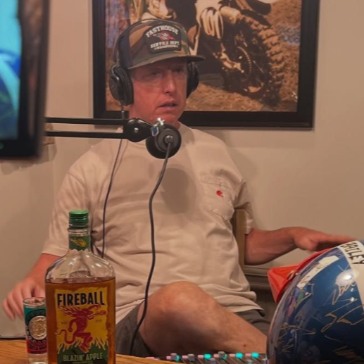 Man wearing headphones sits, speaking into a microphone.  A bottle of Fireball whiskey sits on a table.
