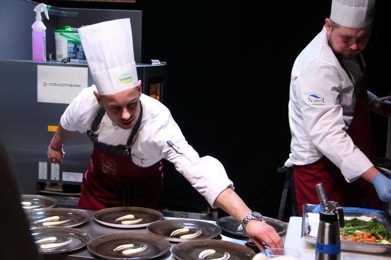 Chefs in white coats and hats plating food in a kitchen with dark background.