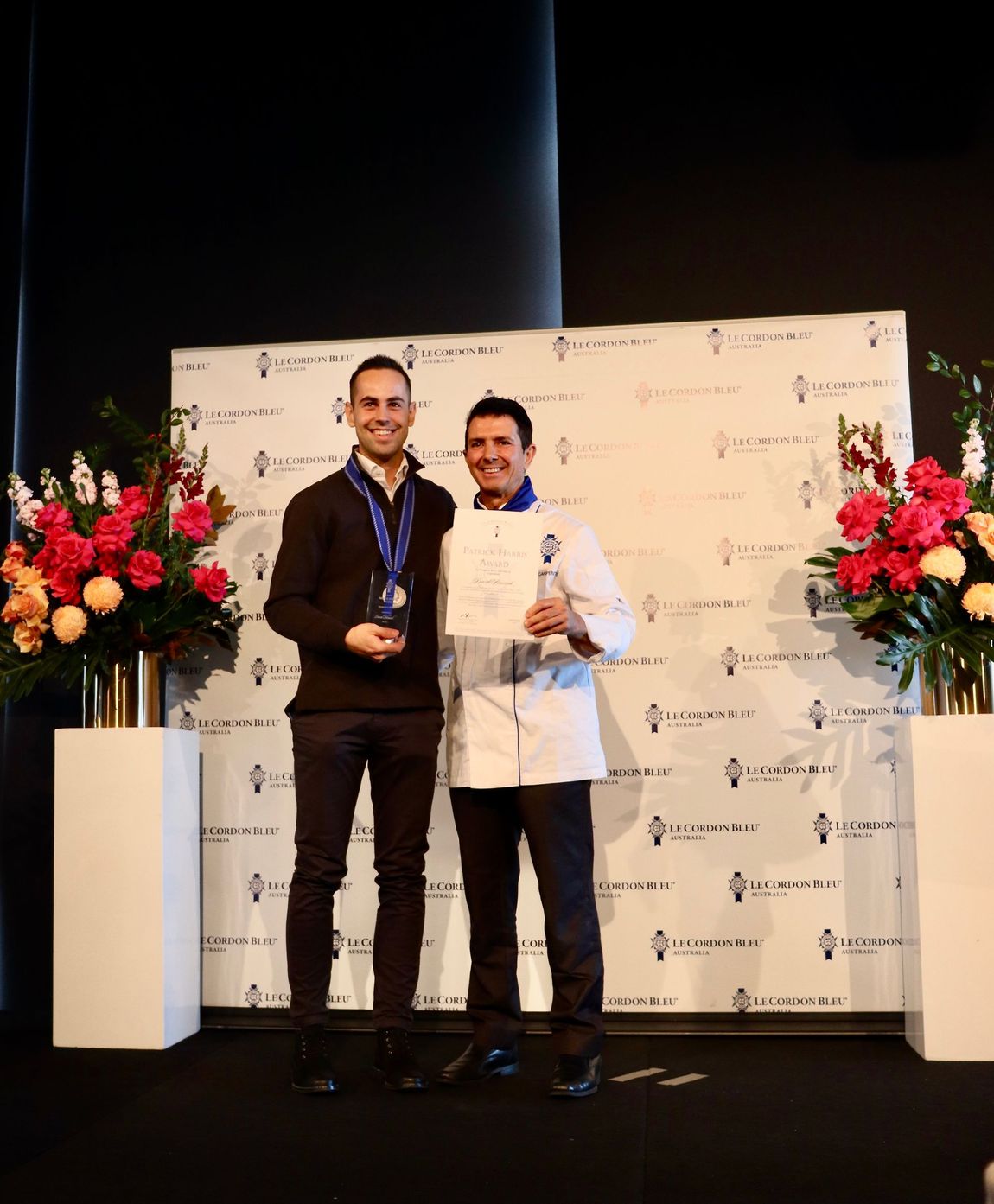 Two men standing in front of a backdrop, one holding a trophy, one holding a certificate.