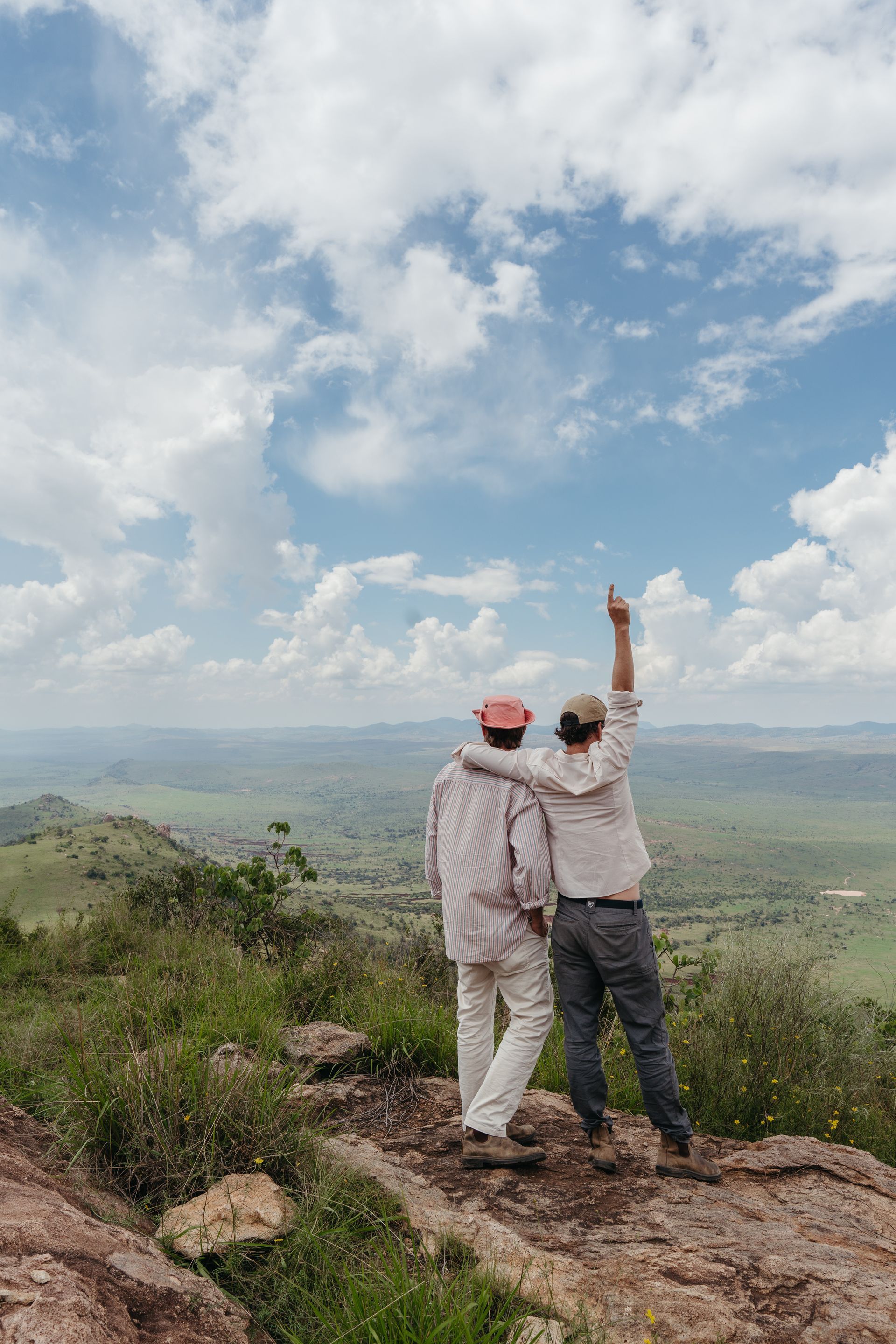 Two people stand on a rocky overlook, raising an arm toward a vast green valley under a cloudy blue sky.