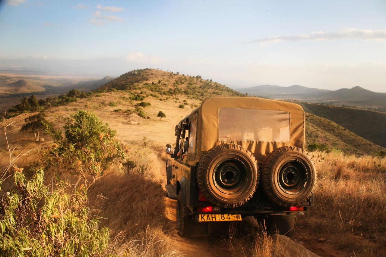 Off-road vehicle driving on a dusty mountain trail at sunset, with spare tires on the back.