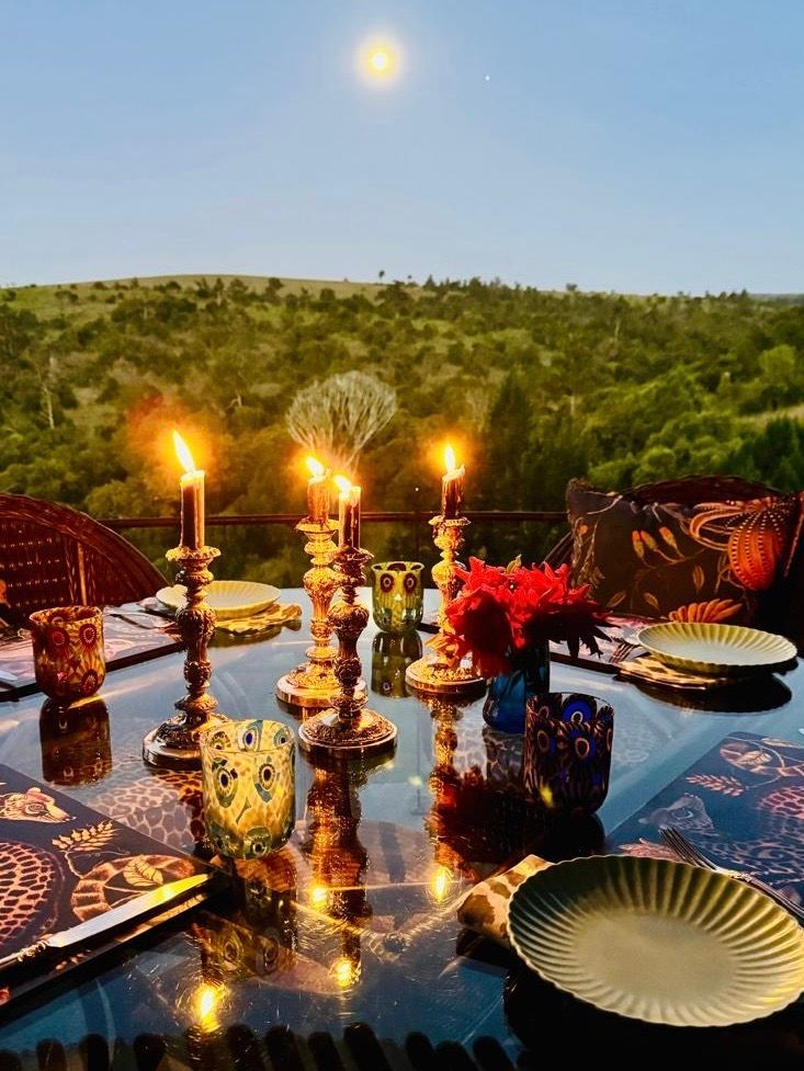 Candlelit outdoor dinner table set with flowers and plates, overlooking green hills under a blue sky