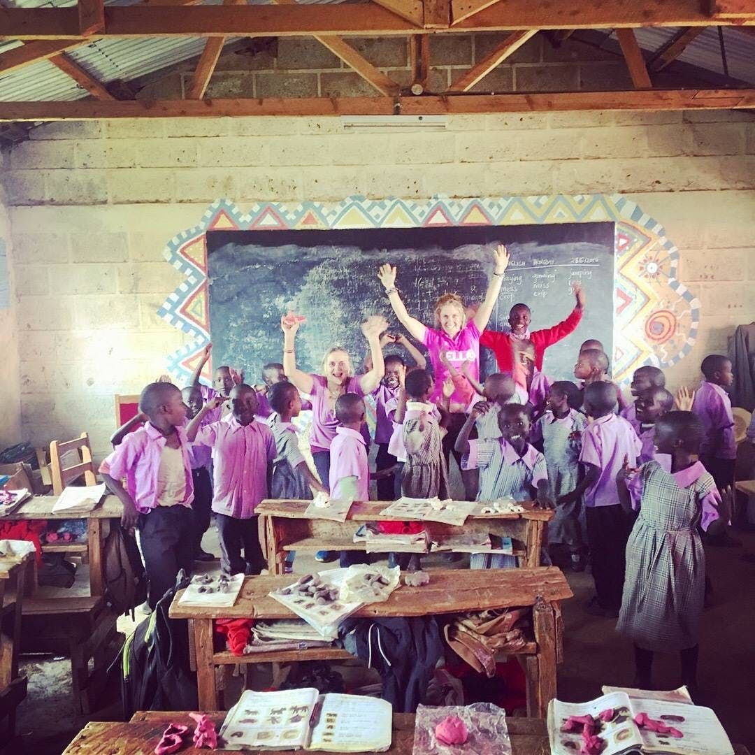 Schoolchildren in pink uniforms pose smiling in a classroom with raised hands and desks in the foreground