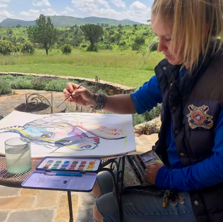 Person painting outdoors at a table with brushes, palette, and water, overlooking green hills.