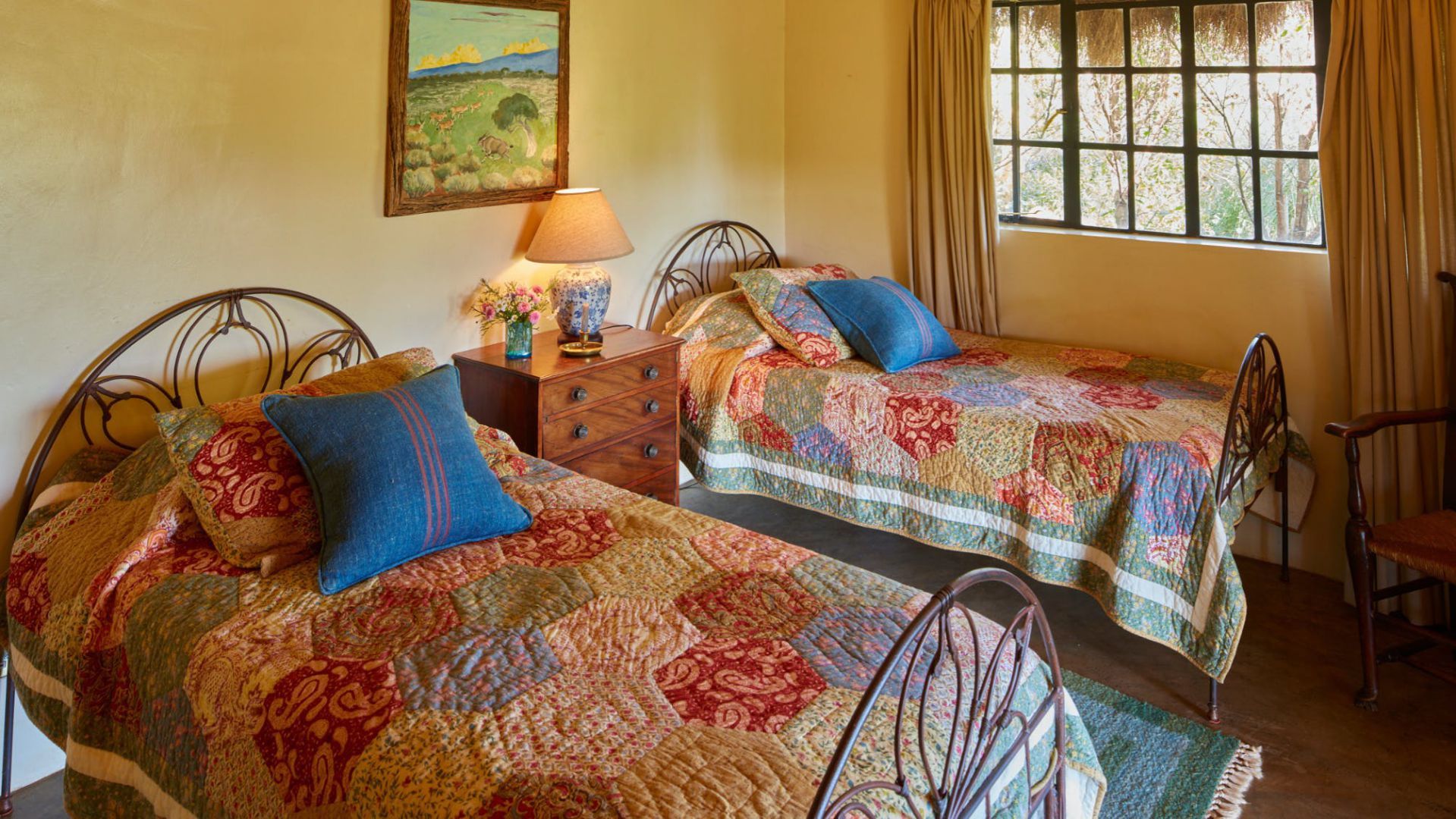 Cozy bedroom with two metal-frame beds, colorful quilts, a bedside lamp, and a window letting in daylight