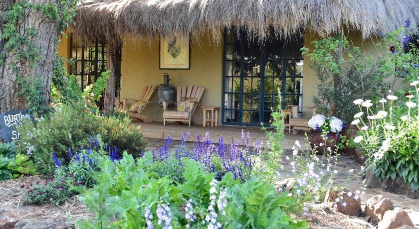 Tropical garden patio with thatched roof, chairs, and lush flowering plants in front.