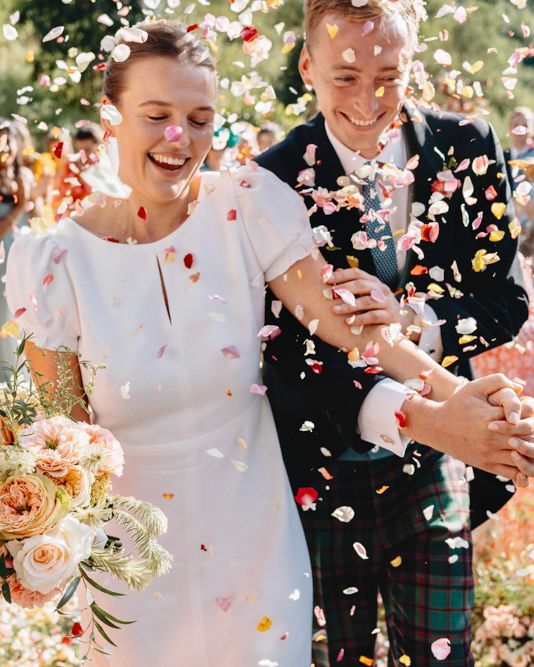 Wedding couple smiling under falling flower petals, holding hands outdoors in sunlight