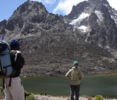 Two hikers stand by a mountain lake beneath steep rocky peaks and patches of snow.