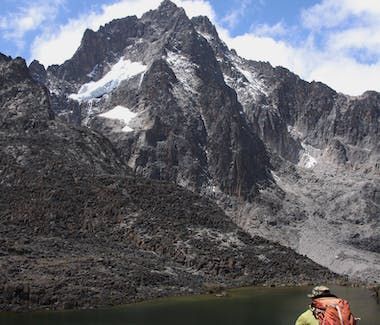 Snow-dusted jagged mountain peaks above a rocky slope, with a small hiker at the lower right.