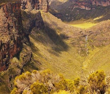 Steep rocky canyon with a sunlit green valley and a thin waterfall in the distance