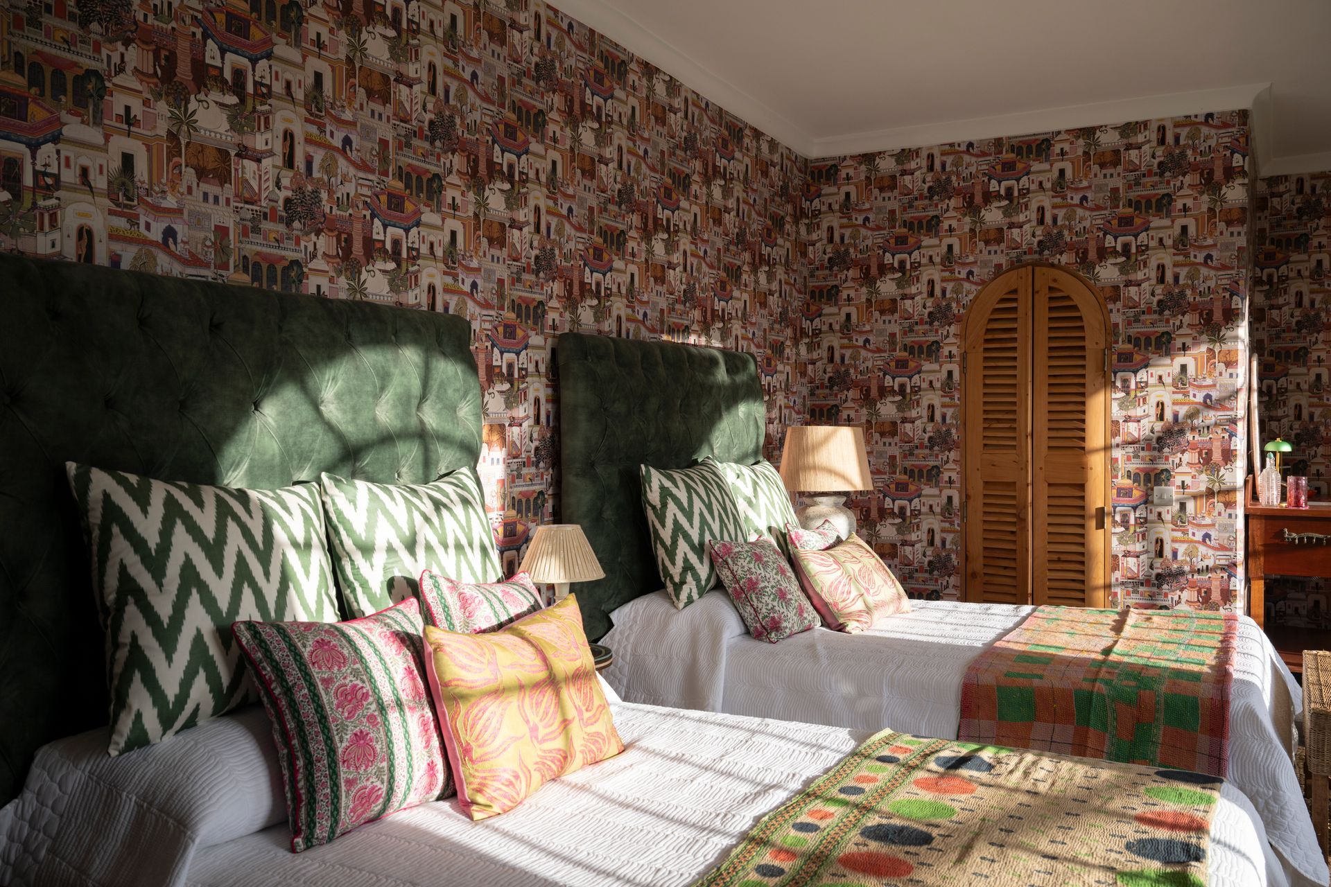 Two neatly made twin beds in a floral wallpapered bedroom with green headboards and sunlight