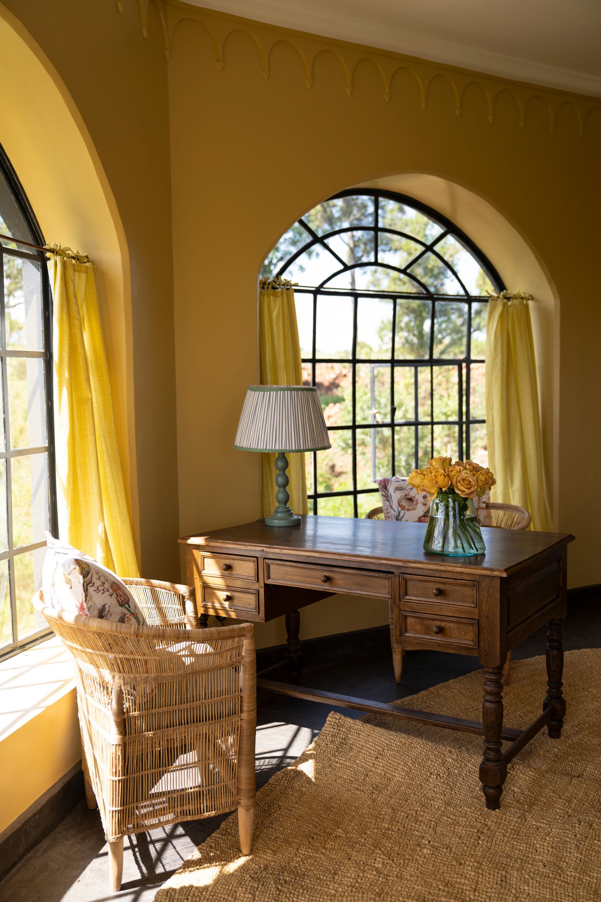 Cozy sunlit room with wicker chairs, wooden desk, lamp, and arched windows with curtains