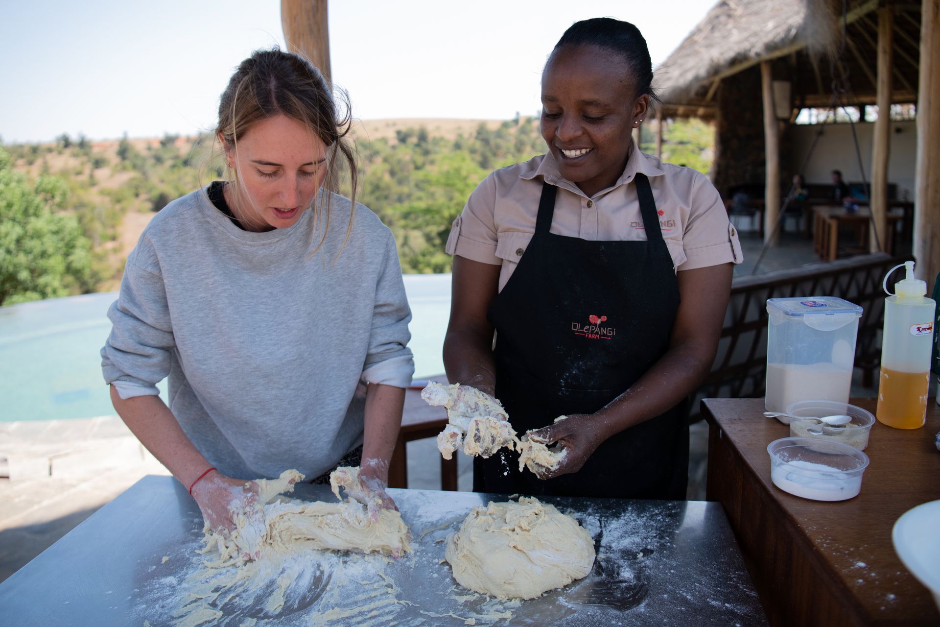 Two people knead dough together at an outdoor table beside a pool.