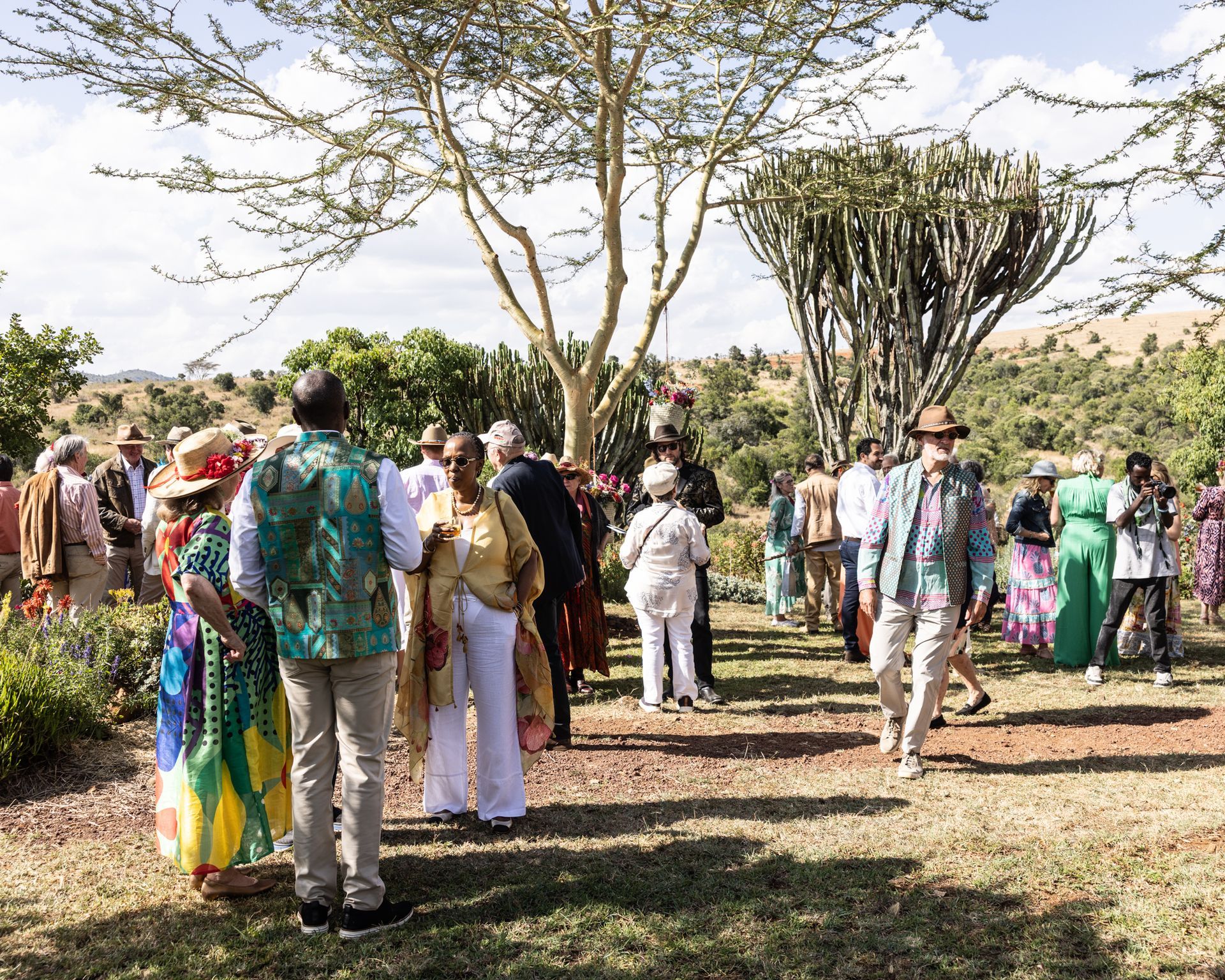 People gathered outdoors under trees, standing in groups on a sunny day.