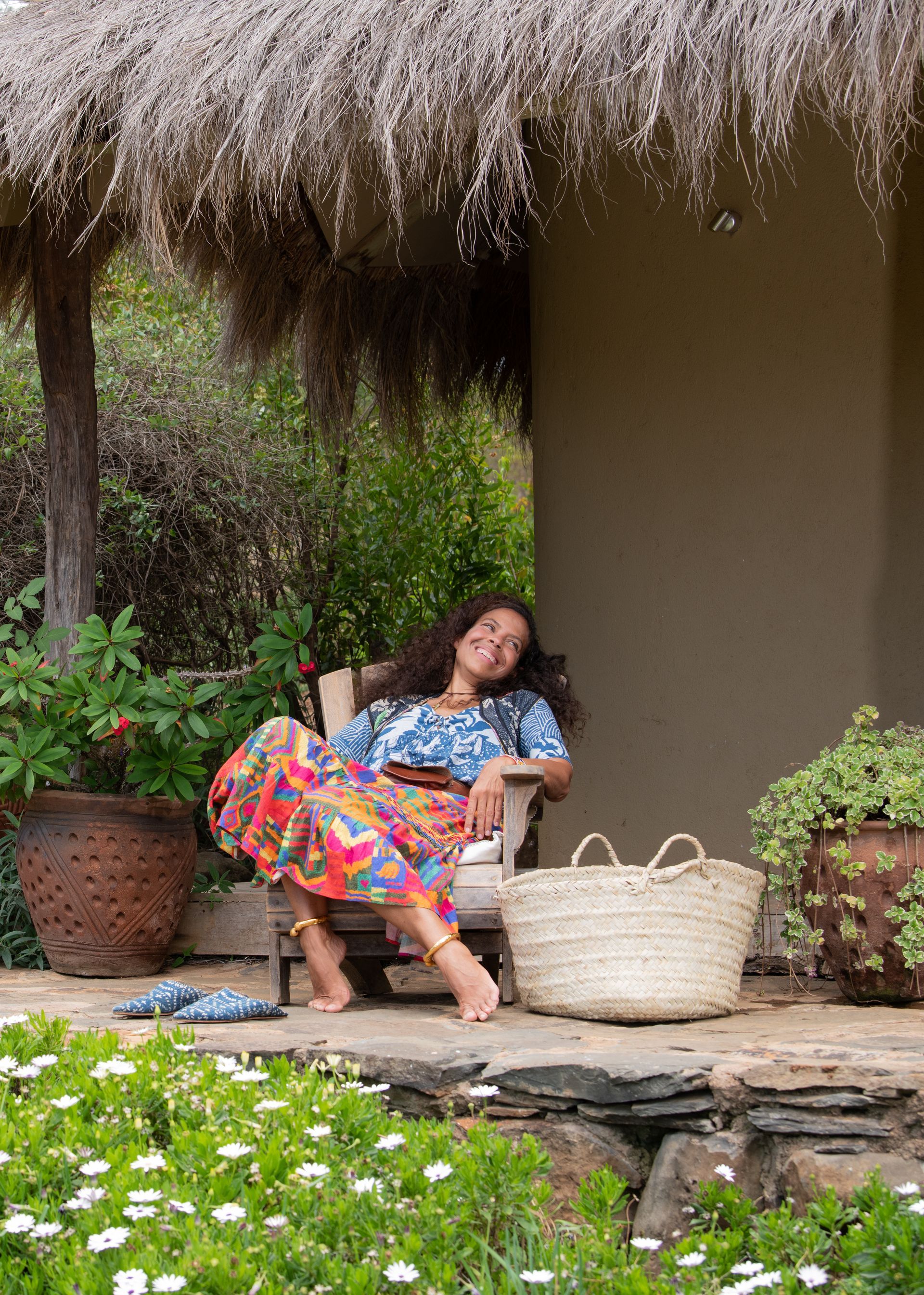 Woman relaxing on a porch in a floral skirt, beside woven baskets and greenery under a thatched roof.
