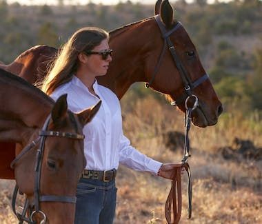 Woman standing beside two saddled horses in a sunlit field, holding one horse’s reins.