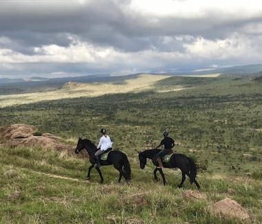 Two riders on black horses overlook a vast green valley under cloudy skies.
