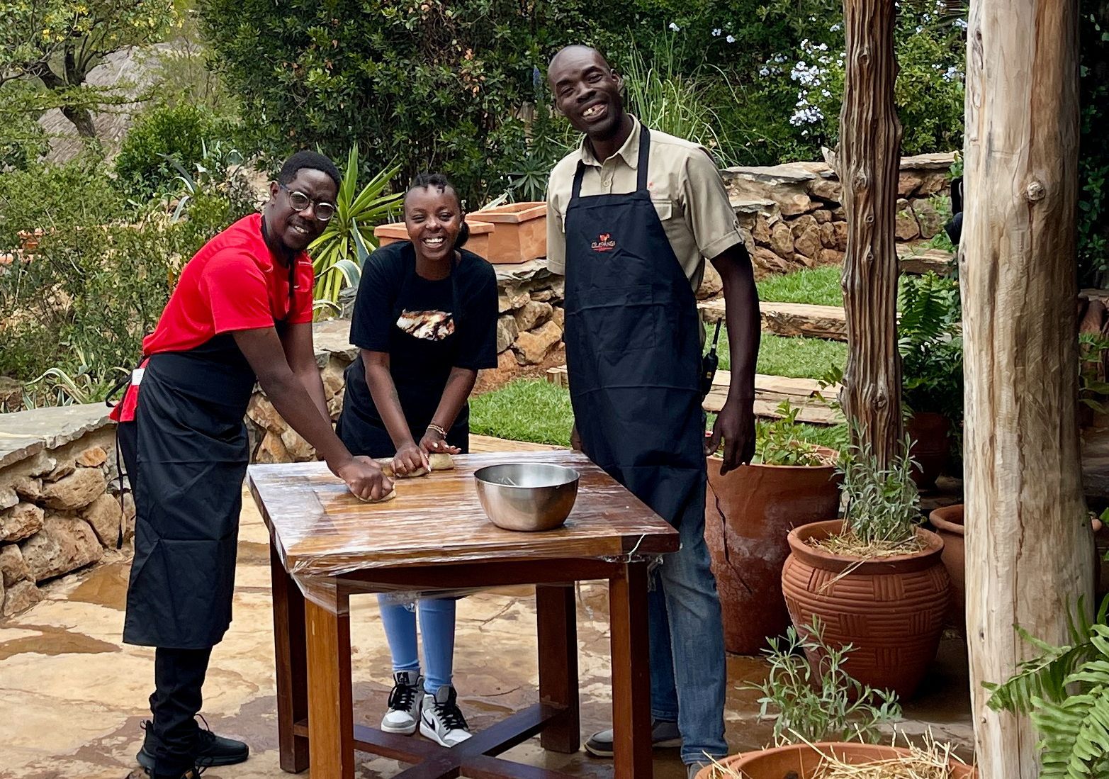 Three people in aprons and gloves stand around a wooden table outdoors, preparing food in a garden.