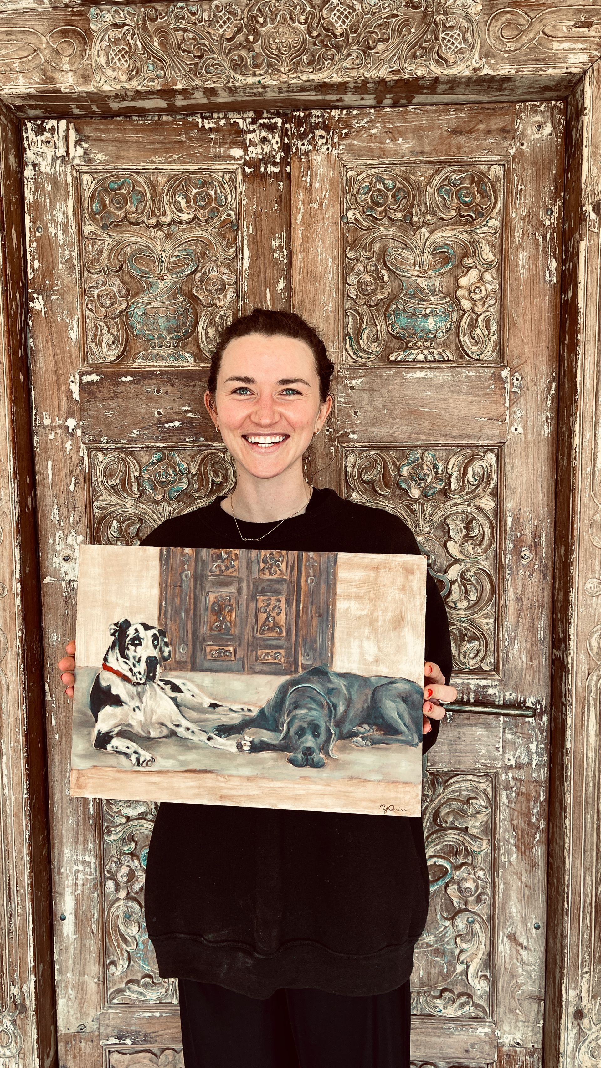 Person smiling and holding a painting in front of a weathered wooden door