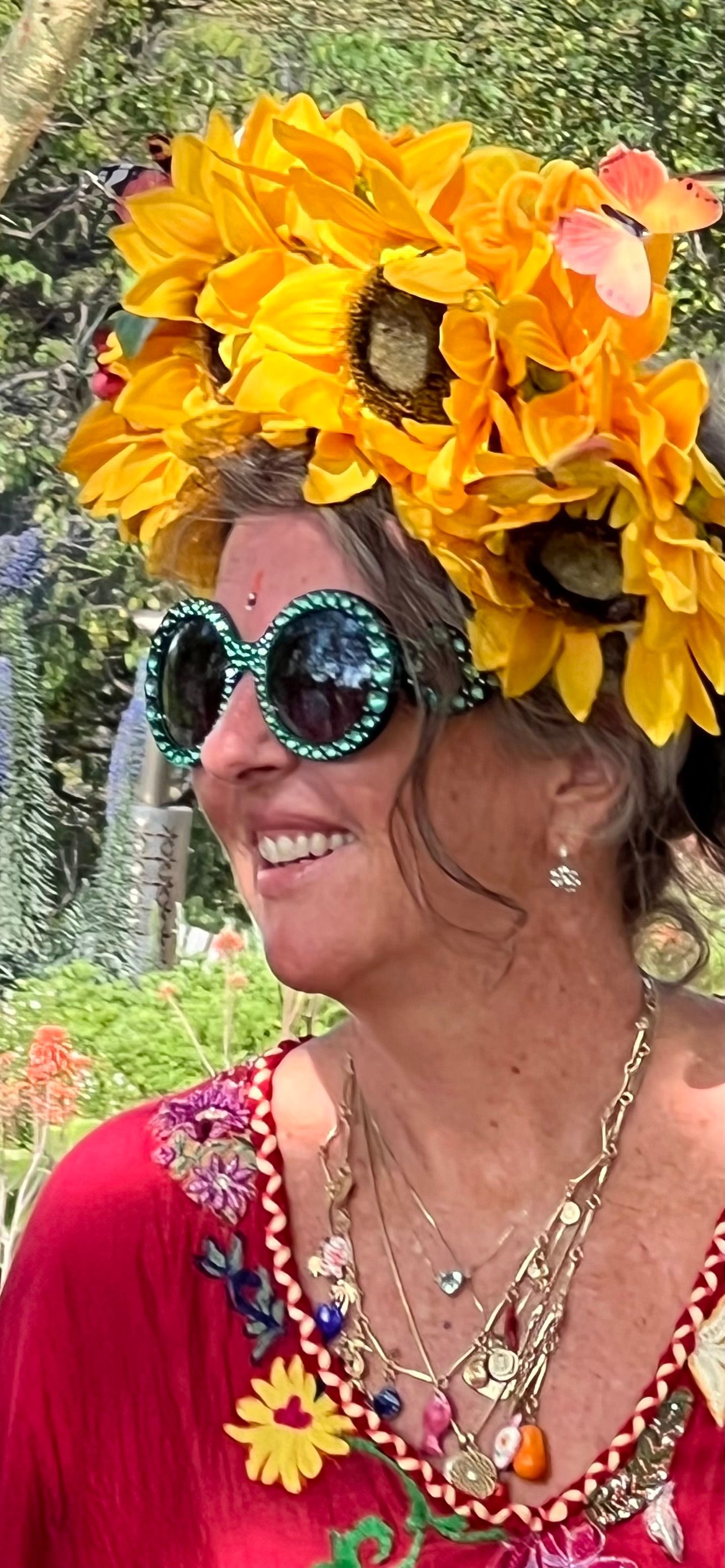 Woman in round sunglasses and a bright yellow flower crown, smiling outdoors in a red floral top.
