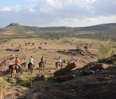 People riding horses across a dry savanna with hills and scattered trees in the distance