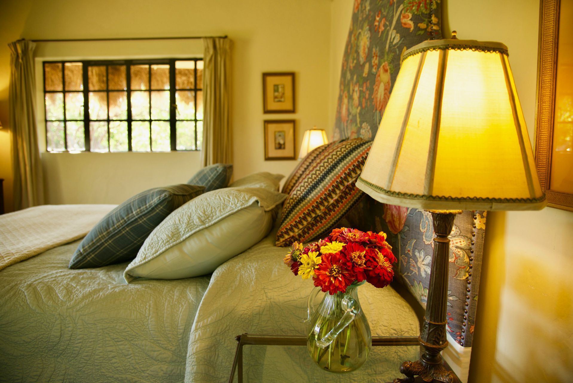 Cozy bedroom with green bedspread, pillows, lamp, and a vase of red flowers by the window