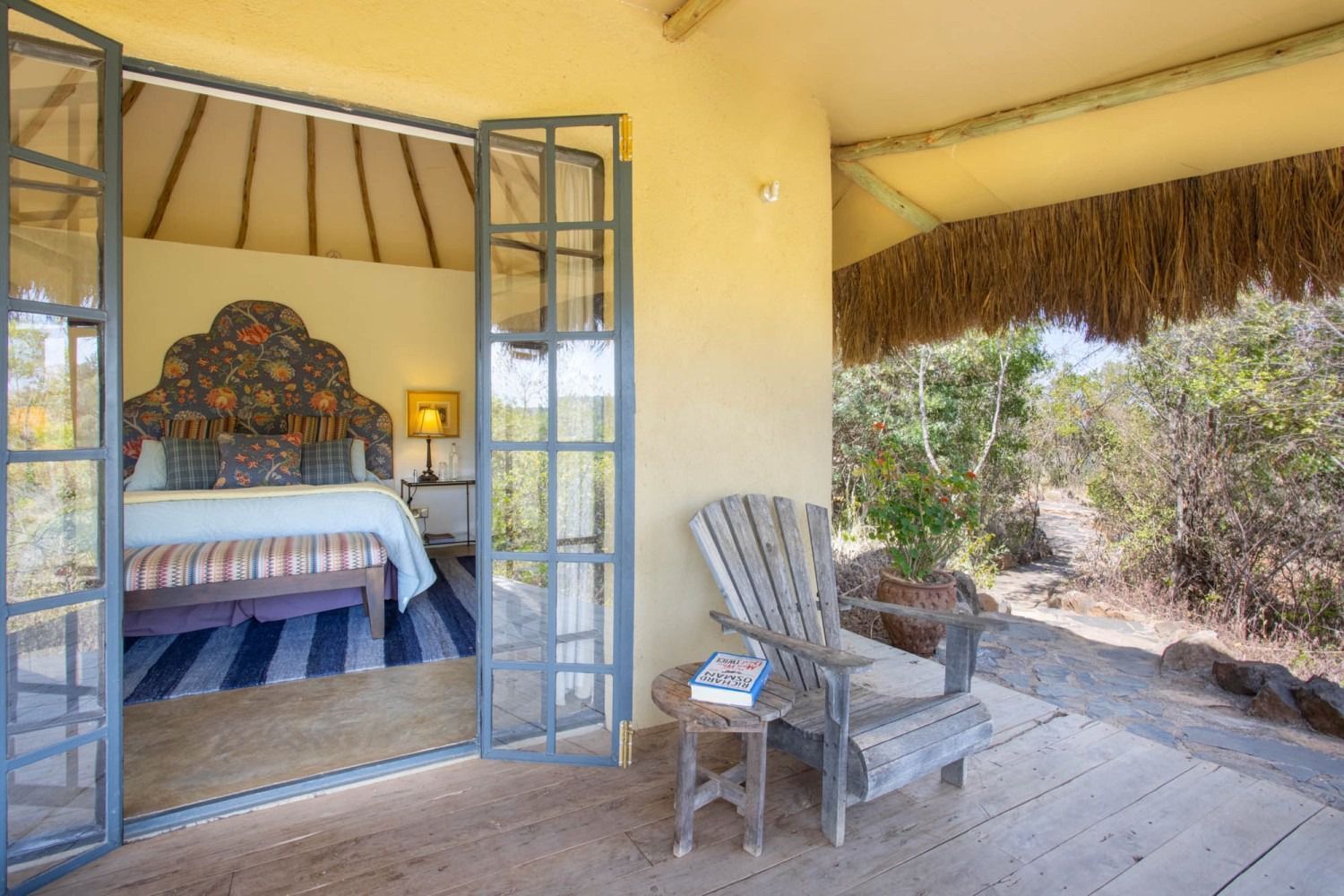 Open-air bedroom with a metal bed, yellow walls, and a shaded porch chair overlooking dry brush