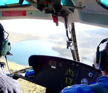 Cockpit view over a blue lake, with two headset-wearing pilots flying above mountains
