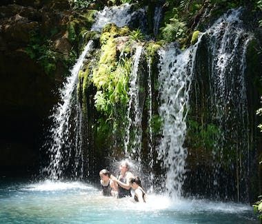 Two people swimming in a turquoise pool beneath a lush waterfall in a forest.