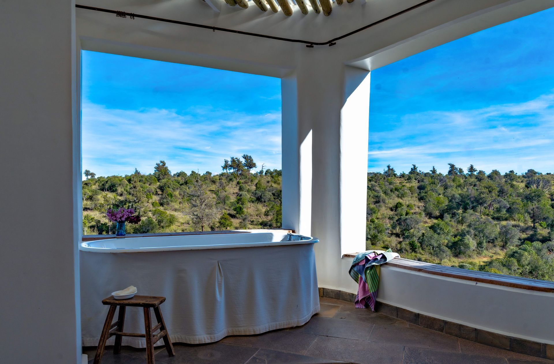 Covered balcony with a stool, draped table, and flower vase overlooking green hills and blue sky