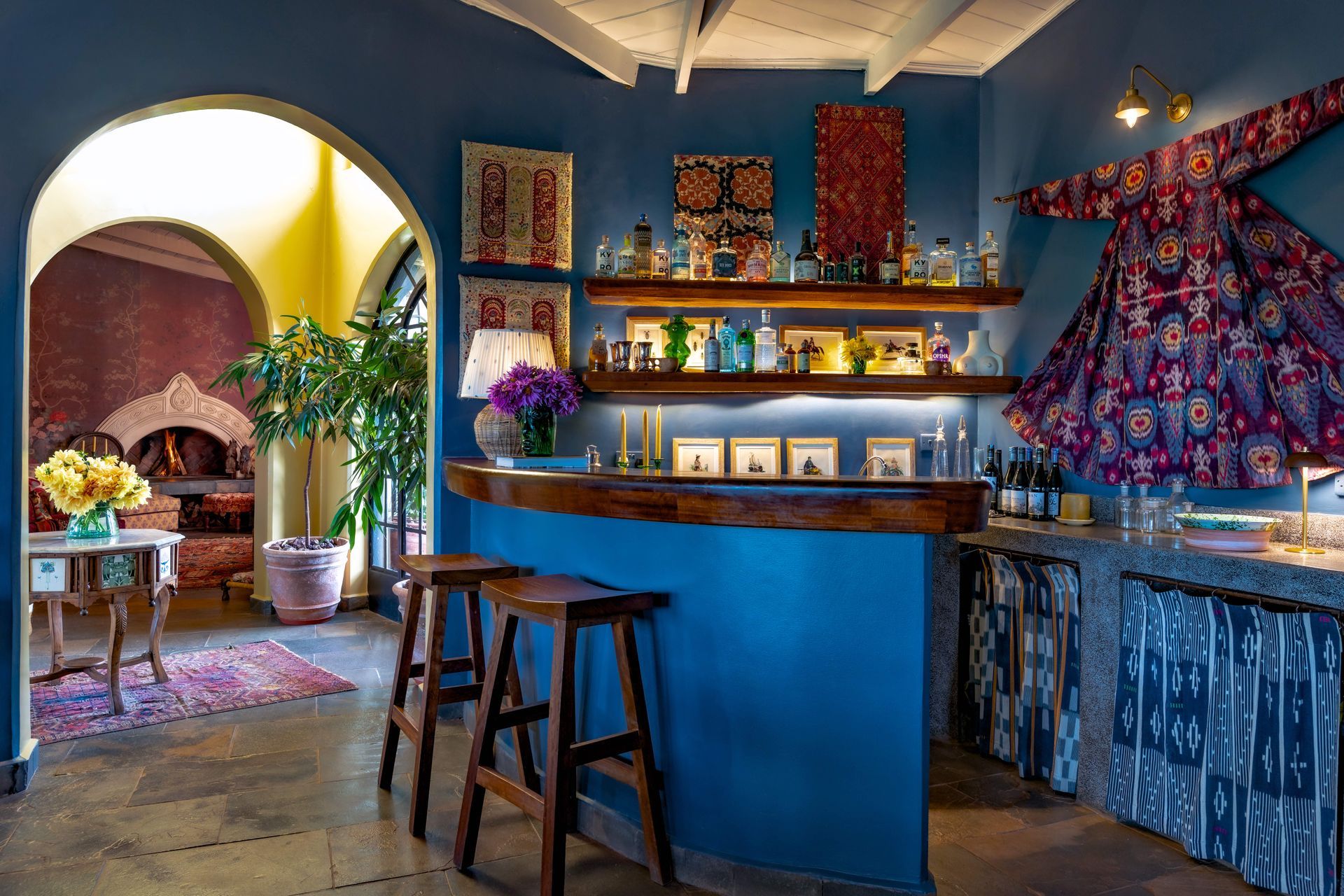 Colorful bohemian kitchen with blue walls, patterned curtains, bar stools, and arched doorway to a cozy adjoining room