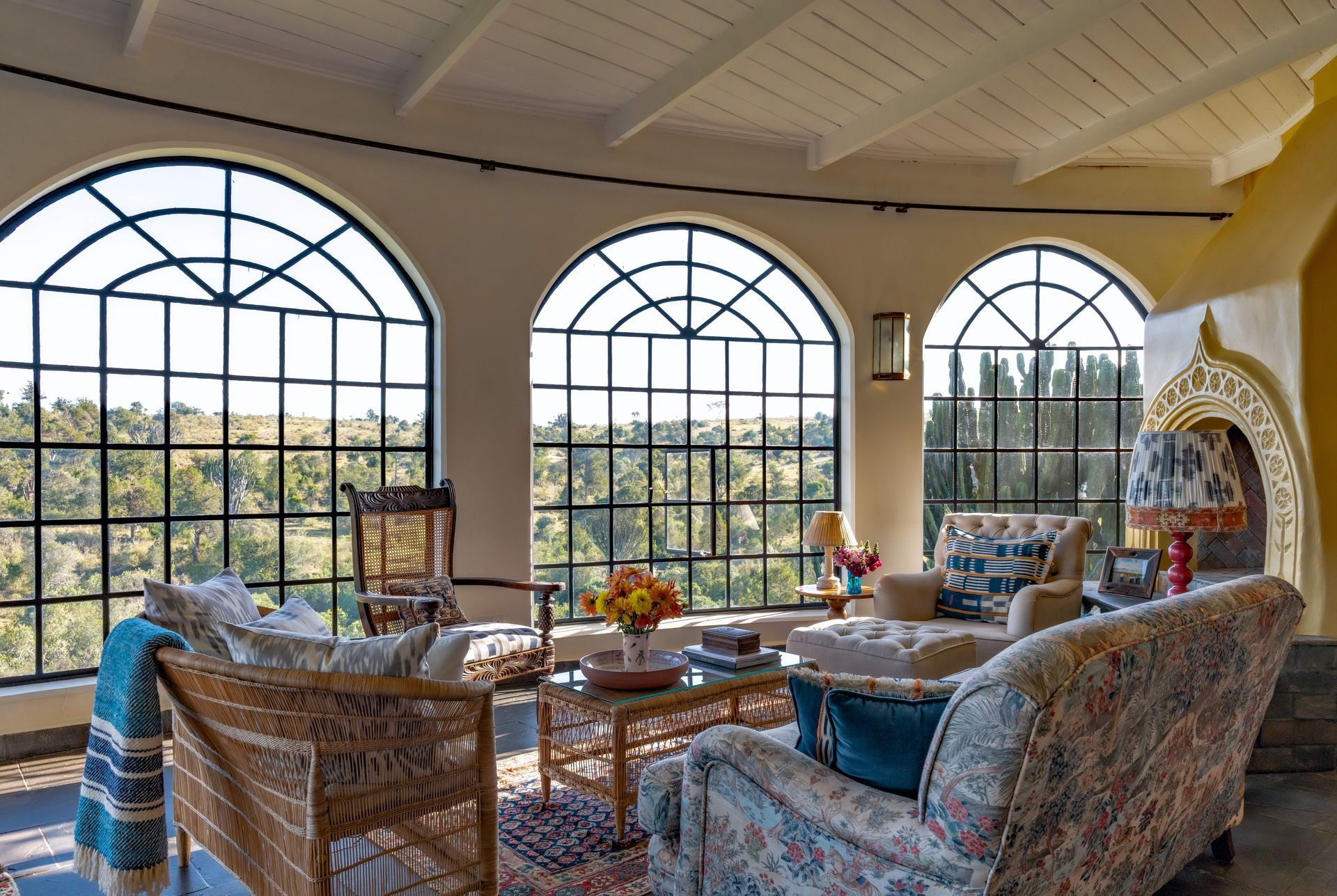 Sunlit sitting room with arched windows, wicker chairs, patterned sofas, and a lace-draped mantel.