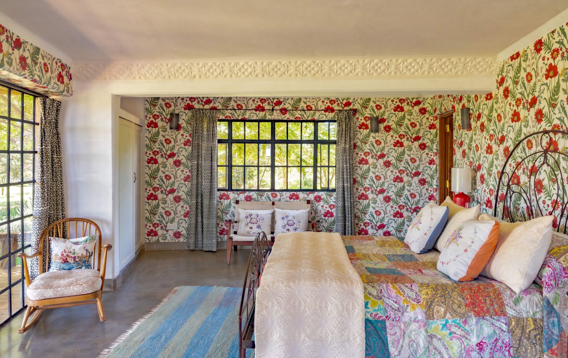 Floral wallpapered bedroom with a bed, wicker chair, and striped rug by large windows