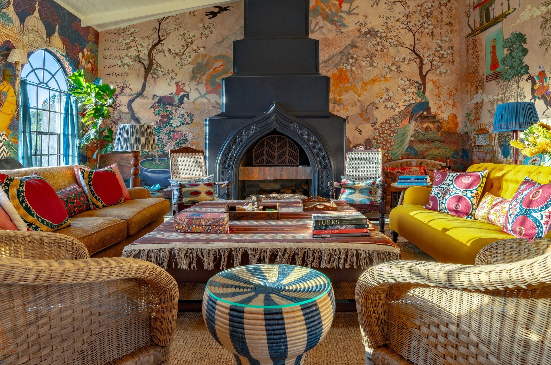 Colorful living room with patterned sofas, woven chairs, and a black fireplace on a mural-covered wall