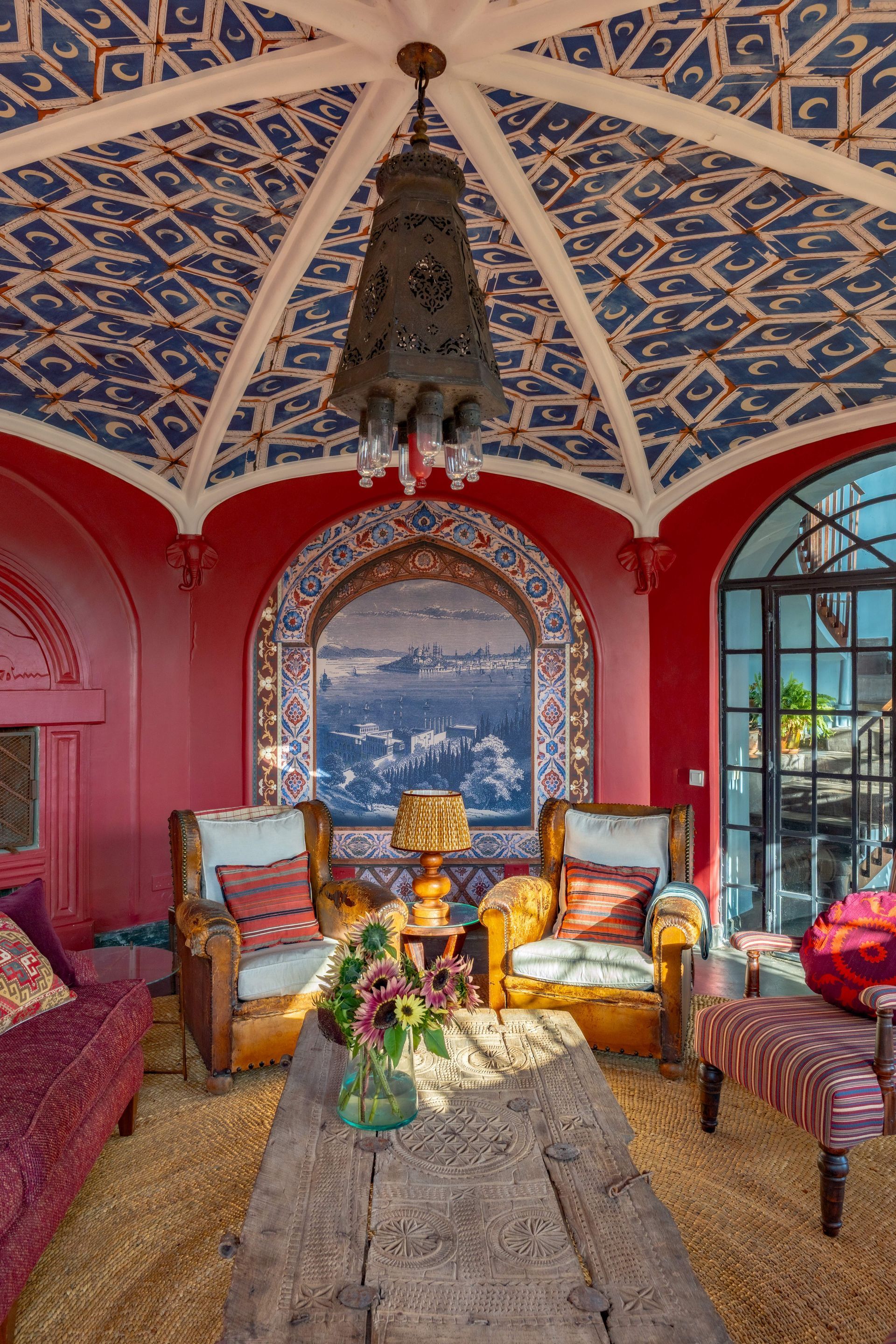 Colorful Moroccan-style lounge with red walls, patterned ceiling, plush seating, and an arched ocean-view window