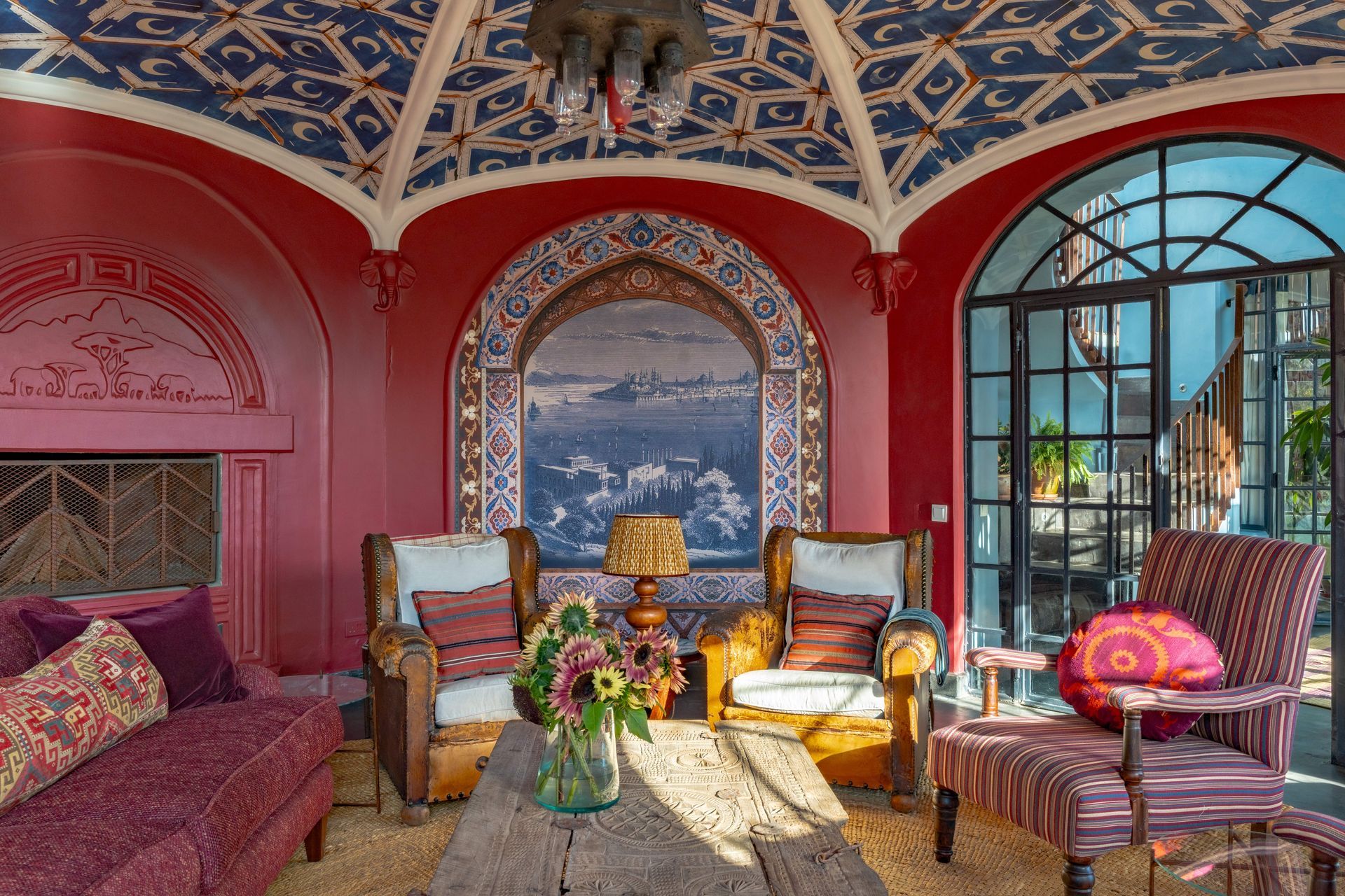 Colorful Moroccan-style lounge with ornate ceiling, red walls, patterned furniture, and a large window view of the sea