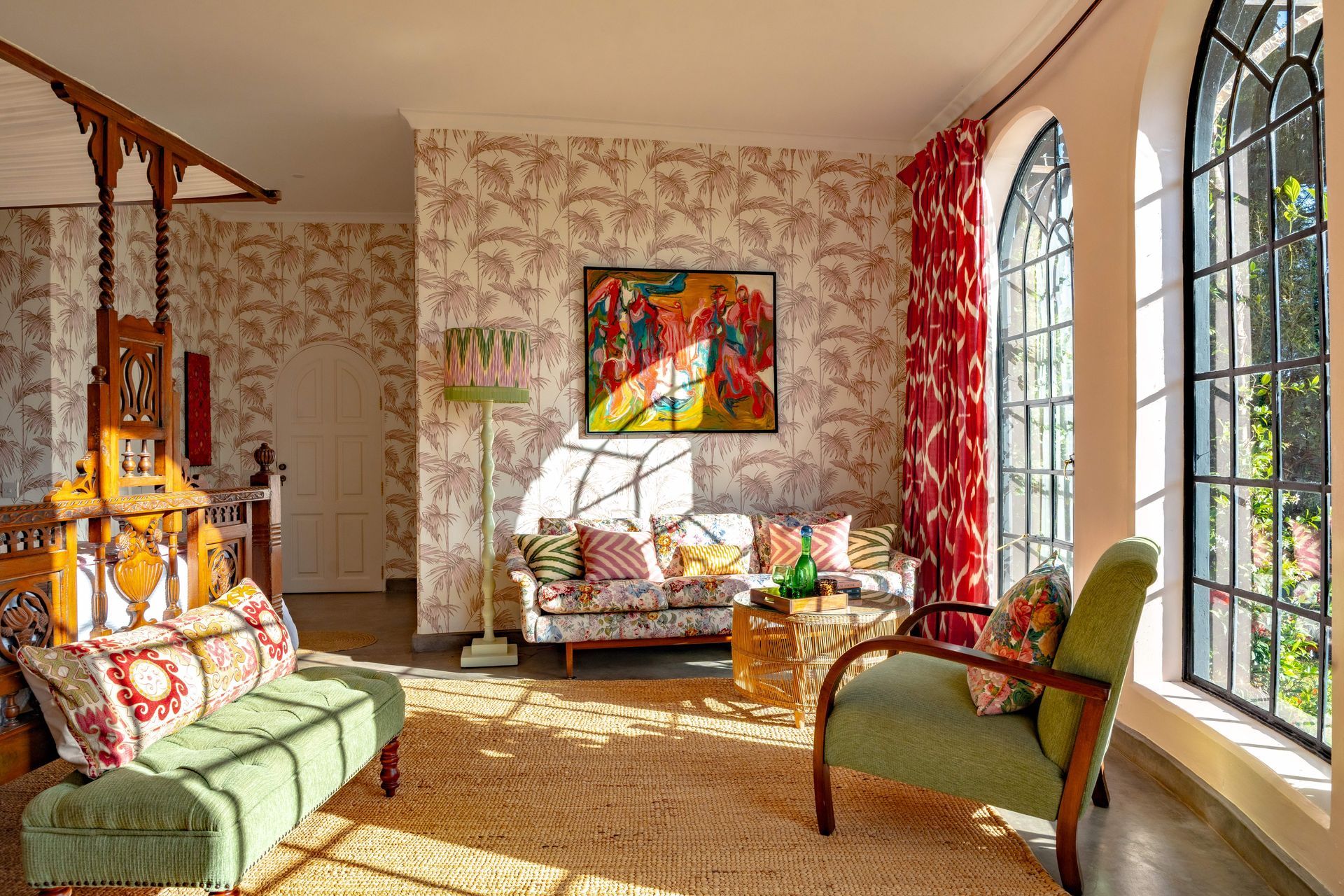 Sunlit living room with floral wallpaper, green sofa, armchairs, colorful art, and tall windows