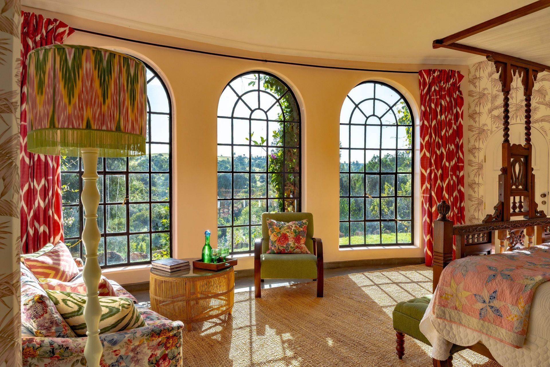 Sunlit room with arched windows, colorful curtains, chairs, and a patterned bedspread.
