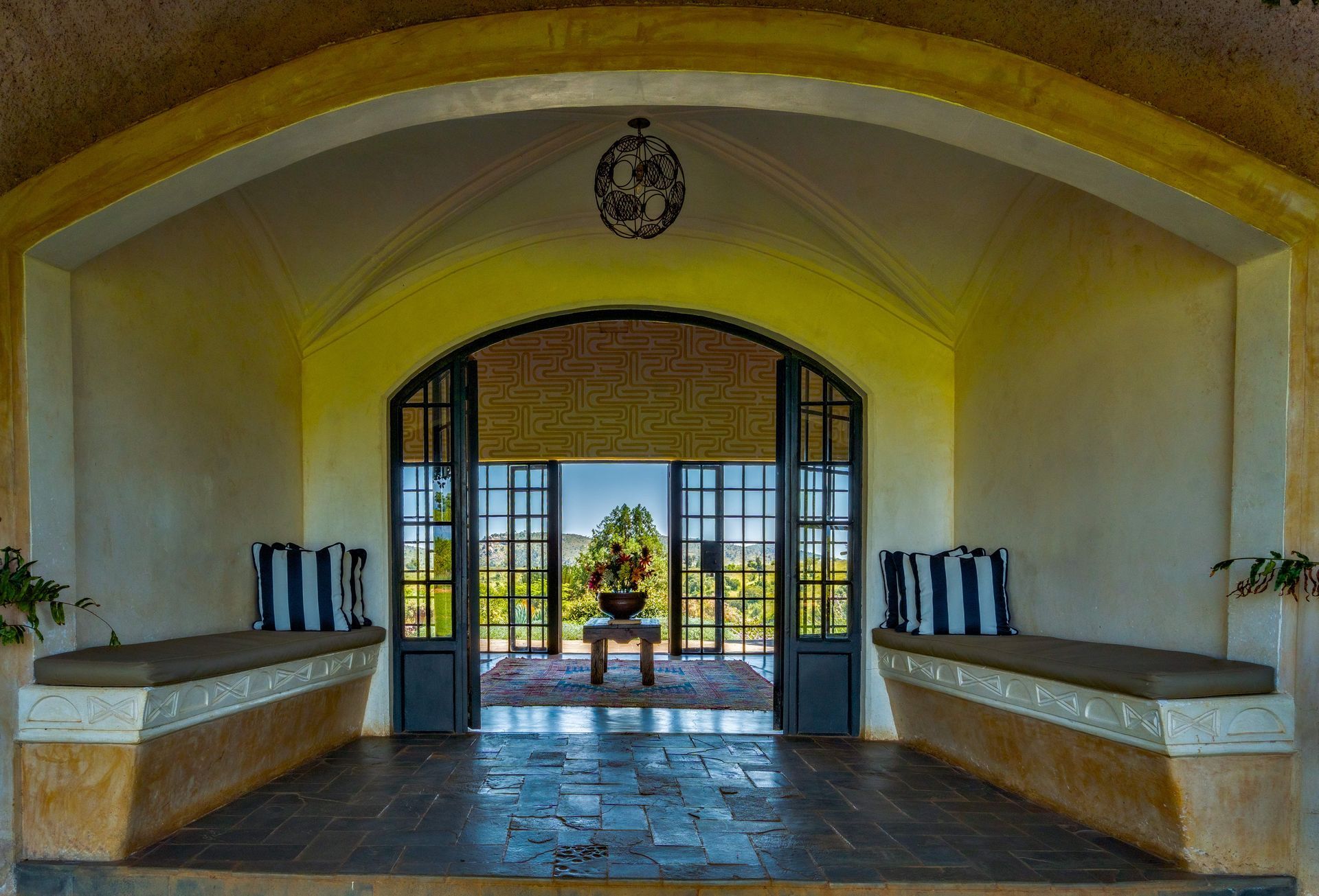 Arched entryway with open double doors framing a sunlit garden and fountain beyond, flanked by benches.